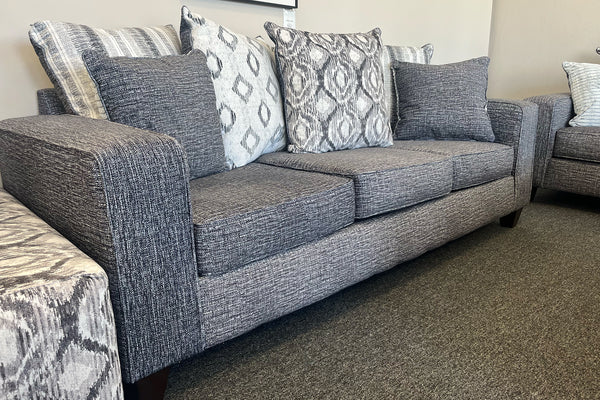 Sofá moderno tapizado en tela gris jaspeada, con cojines decorativos en patrones geométricos y texturas en tonos gris, blanco y negro, exhibido en una tienda de muebles.