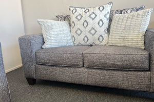 Loveseat moderno tapizado en tela gris jaspeada, con cojines decorativos en patrones geométricos y texturas en tonos gris, blanco y negro, exhibido en una tienda de muebles.