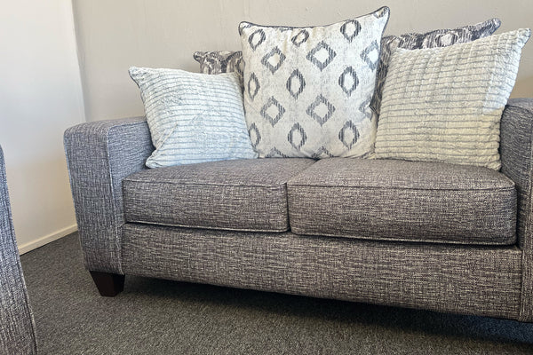 Loveseat moderno tapizado en tela gris jaspeada, con cojines decorativos en patrones geométricos y texturas en tonos gris, blanco y negro, exhibido en una tienda de muebles.