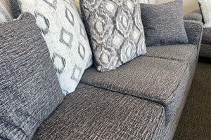 Primer plano de sofá tapizado en tela gris jaspeada con cojines decorativos de patrones geométricos en tonos gris, blanco y negro, exhibido en una sala de muebles.