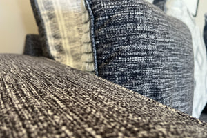 Primer plano de sofá tapizado en tela gris jaspeada con textura tejida visible y cojines decorativos en tonos gris, blanco y negro, exhibido en una tienda de muebles.