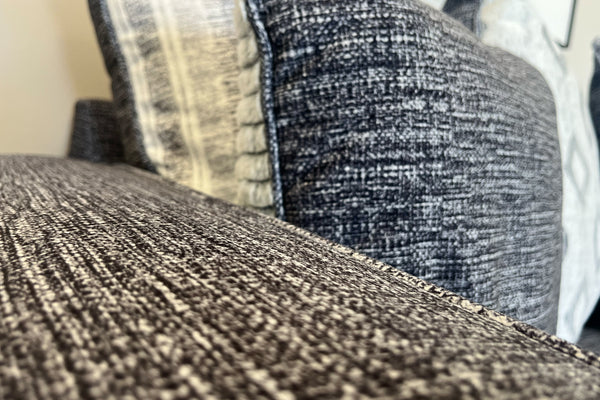 Primer plano de sofá tapizado en tela gris jaspeada con textura tejida visible y cojines decorativos en tonos gris, blanco y negro, exhibido en una tienda de muebles.