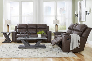 Sofá y loveseat reclinable de color marrón oscuro con objetos decorativos en una sala neutral con una ventana en el fondo. 