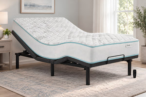 Una base de cama eléctrica ajustable con un mattress acolchado blanco, presentada en un dormitorio luminoso y moderno con una alfombra gris, una mesita de noche y una ventana.