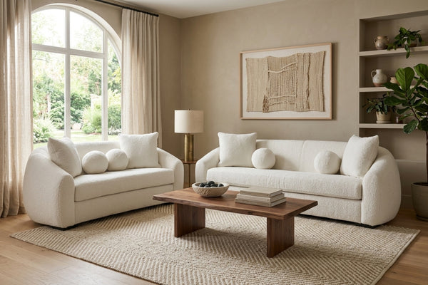 Sala contemporánea con sofá y loveseat curvos en color crema, cojines decorativos redondos y mesa de centro de madera sobre alfombra beige, en un espacio luminoso y minimalista.