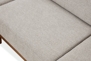 Primer plano del cojín de un sofá tapizado en tela texturizada de color gris claro, con una pequeña parte de una pata de madera marrón visible en la esquina inferior izquierda.