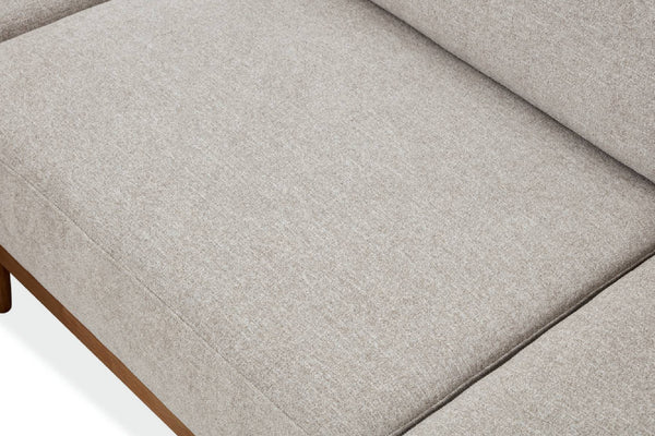 Primer plano del cojín de un sofá tapizado en tela texturizada de color gris claro, con una pequeña parte de una pata de madera marrón visible en la esquina inferior izquierda.