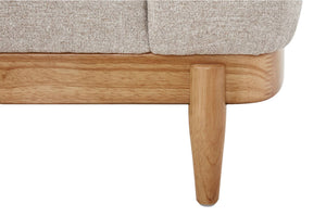 Primer plano de la esquina de un sofá o cama con tapizado texturizado en beige claro, estructura de madera natural y pata de madera cónica sobre un fondo blanco.
