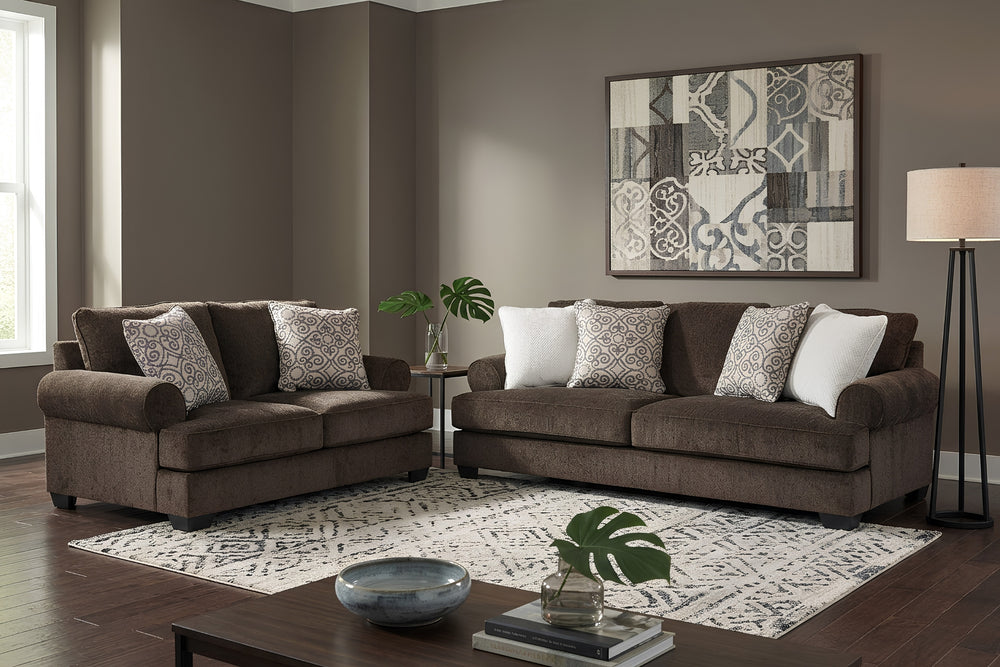 Sofá y loveseat de color marrón con cojines decorativos sobre fondo blanco. 