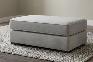 Otoman de color gris claro de forma rectangular con patas cuadradas y bajitas en una sala neutral con alfombra gris. 
