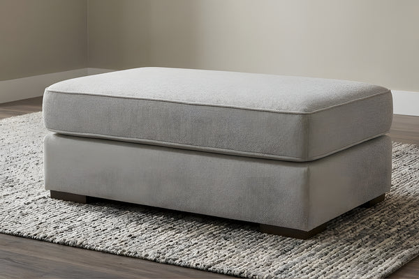 Otoman de color gris claro de forma rectangular con patas cuadradas y bajitas en una sala neutral con alfombra gris. 