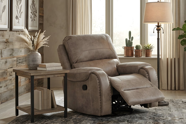 Butaca reclinable individual en color gris taupé con reposapiés extendido, brazos acolchados con detalles de tachuelas decorativas y respaldo amplio, ubicado junto a una mesa auxiliar y lámpara de pie en una sala de estar moderna.