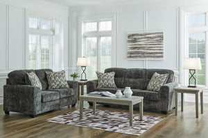 Sofá y loveseat de color gris oscuro con cojines decorativos en una sala moderna con ventanas. 