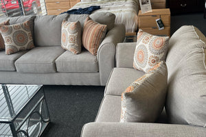 Juego de sala con sofá y loveseat tapizados en tela beige, decorados con cojines estampados en tonos tierra, exhibidos en una tienda de muebles junto a una mesa de centro de aluminio y vidrio.