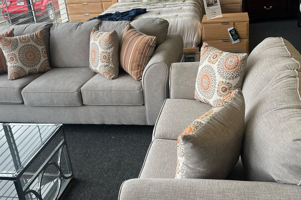 Juego de sala con sofá y loveseat tapizados en tela beige, decorados con cojines estampados en tonos tierra, exhibidos en una tienda de muebles junto a una mesa de centro de aluminio y vidrio.