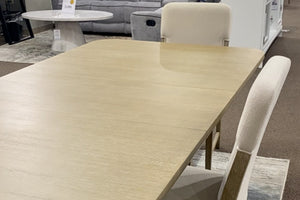 Vista cercana de mesa de comedor rectangular en acabado madera clara con superficie amplia y lisa, acompañada por sillas tapizadas en tela color marfil. El conjunto exhibe un diseño contemporáneo de líneas limpias y tonos neutros.