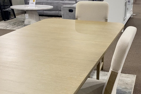 Vista cercana de mesa de comedor rectangular en acabado madera clara con superficie amplia y lisa, acompañada por sillas tapizadas en tela color marfil. El conjunto exhibe un diseño contemporáneo de líneas limpias y tonos neutros.