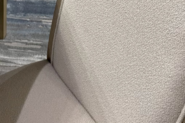 Primer plano de silla de comedor tapizada en tela color marfil con textura bouclé, mostrando el respaldo y asiento acolchados. La imagen resalta el tejido suave, los acabados y el diseño elegante de estilo contemporáneo.