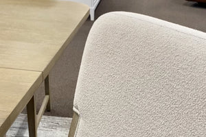 Primer plano de silla de comedor tapizada en tela color marfil con textura tipo bouclé, junto a una mesa rectangular en acabado madera clara. La imagen destaca los materiales, acabados suaves y el diseño contemporáneo del conjunto.