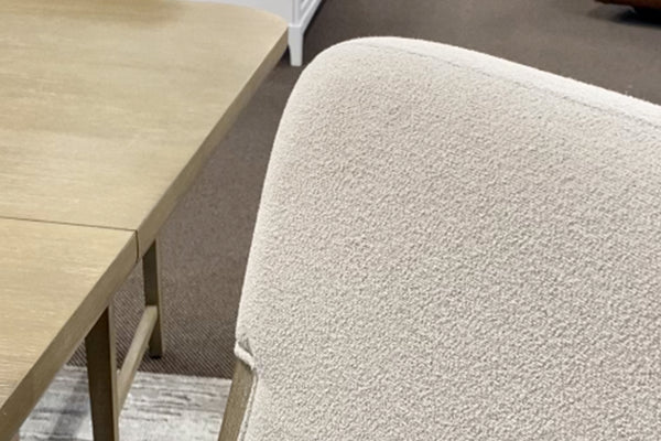 Primer plano de silla de comedor tapizada en tela color marfil con textura tipo bouclé, junto a una mesa rectangular en acabado madera clara. La imagen destaca los materiales, acabados suaves y el diseño contemporáneo del conjunto.
