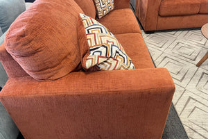 Sofá naranja con un cojín estampado sobre un suelo alfombrado en una sala de exposición.
