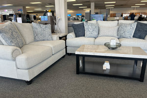 Juego de sala con loveseat y sofá tapizados en tela beige clara, decorados con cojines en tonos azul y gris, acompañado de mesa de centro rectangular con cubierta tipo mármol en una sala de exhibición de muebles.