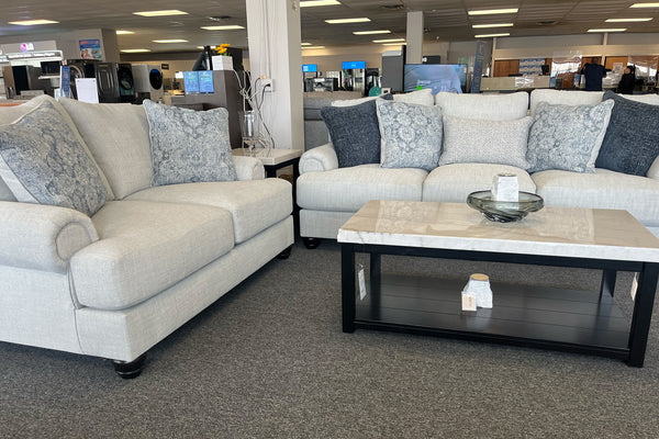 Juego de sala con loveseat y sofá tapizados en tela beige clara, decorados con cojines en tonos azul y gris, acompañado de mesa de centro rectangular con cubierta tipo mármol en una sala de exhibición de muebles.