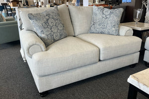 Loveseat tapizados en tela beige clara, decorados con cojines en tonos azul y gris, en una sala de exhibición de muebles.