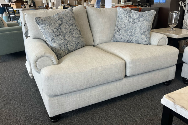 Loveseat tapizados en tela beige clara, decorados con cojines en tonos azul y gris, en una sala de exhibición de muebles.