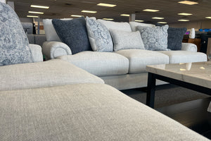 Sala de exhibición con sofá y loveseat tapizados en tela beige clara, cojines decorativos en tonos azul, gris y blanco, y mesa de centro rectangular con cubierta tipo mármol y base negra.
