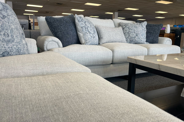 Sala de exhibición con sofá y loveseat tapizados en tela beige clara, cojines decorativos en tonos azul, gris y blanco, y mesa de centro rectangular con cubierta tipo mármol y base negra.