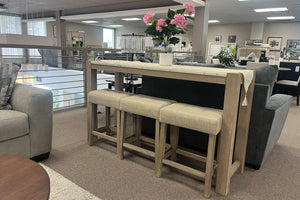 Mesa alta rectangular de madera clara con tres bancos tapizados en color beige, exhibida en un showroom de muebles modernos. La mesa está decorada con un camino de mesa y un arreglo floral rosa, rodeada de sofás y mobiliario contemporáneo en un espacio abierto y luminoso.