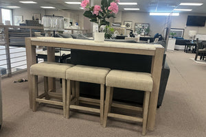 Mesa alta rectangular de madera clara con tres bancos tapizados en color beige, exhibida en un showroom de muebles modernos. La mesa está decorada con un camino de mesa y un arreglo floral rosa, rodeada de sofás y mobiliario contemporáneo en un espacio abierto y luminoso.