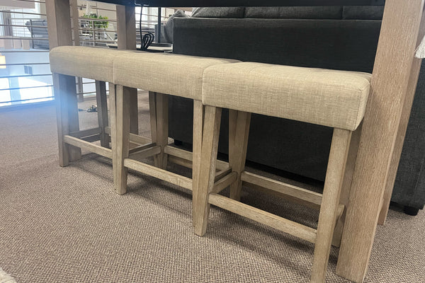 Tres bancos altos con estructura de madera clara y asientos tapizados en tela beige, colocados debajo de una mesa alta rectangular en un showroom de muebles contemporáneos. El diseño incluye líneas rectas y reposapiés integrados para mayor comodidad.