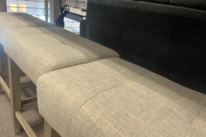Primer plano de bancos altos con asientos acolchados tapizados en tela beige y estructura de madera clara. La imagen destaca la textura de la tela y el diseño moderno de los bancos colocados junto a una mesa alta en un showroom de muebles contemporáneos.