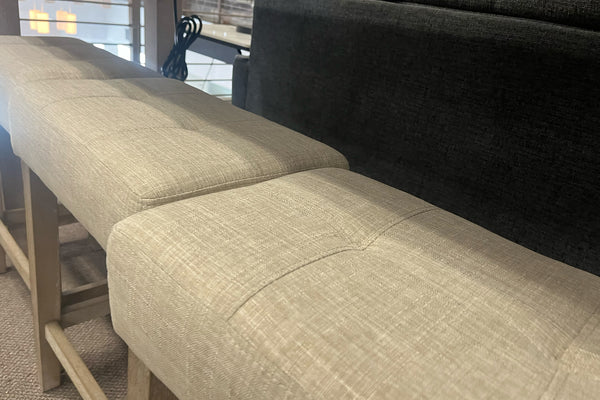 Primer plano de bancos altos con asientos acolchados tapizados en tela beige y estructura de madera clara. La imagen destaca la textura de la tela y el diseño moderno de los bancos colocados junto a una mesa alta en un showroom de muebles contemporáneos.
