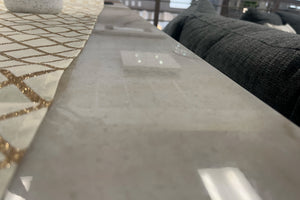 Primer plano de una mesa alta con superficie lisa en acabado tipo mármol claro y camino de mesa decorativo con patrón geométrico dorado. Al fondo se observa parcialmente un sofá gris oscuro en una sala de exhibición de muebles modernos.