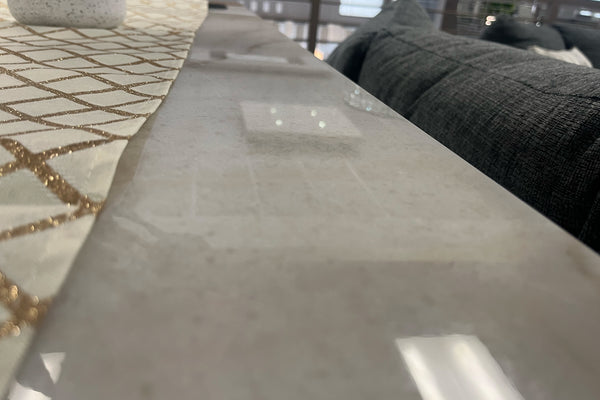 Primer plano de una mesa alta con superficie lisa en acabado tipo mármol claro y camino de mesa decorativo con patrón geométrico dorado. Al fondo se observa parcialmente un sofá gris oscuro en una sala de exhibición de muebles modernos.