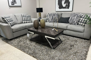 Sala moderna con sofá y loveseat tapizados en tela gris claro, cojines decorativos geométricos en tonos gris y blanco, mesa de centro de madera oscura con detalles metálicos y alfombra gris de textura suave.