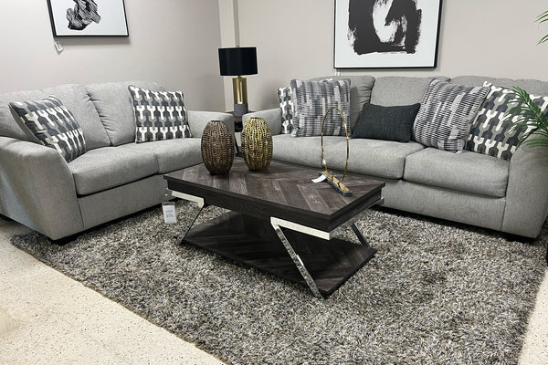 Sala moderna con sofá y loveseat tapizados en tela gris claro, cojines decorativos geométricos en tonos gris y blanco, mesa de centro de madera oscura con detalles metálicos y alfombra gris de textura suave.