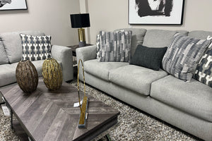 Sala moderna con sofá y loveseat tapizados en tela gris claro, cojines decorativos geométricos en tonos gris y blanco, mesa de centro de madera oscura con detalles metálicos y alfombra gris de textura suave.