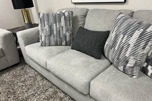 Sofá tapizado en tela gris claro con cojines decorativos en patrones geométricos blanco y negro, acompañado de cojín lumbar oscuro y alfombra gris de textura suave en una sala de exhibición moderna.