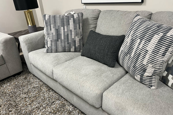 Sofá tapizado en tela gris claro con cojines decorativos en patrones geométricos blanco y negro, acompañado de cojín lumbar oscuro y alfombra gris de textura suave en una sala de exhibición moderna.