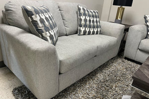 Loveseat tapizado en tela gris claro con cojines decorativos geométricos en tonos gris y blanco, ubicado sobre una alfombra gris de textura suave en una sala de exhibición moderna.