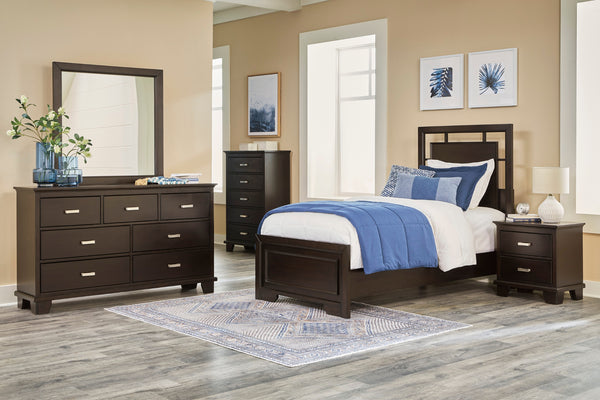 Conjunto de dormitorio con gavetero con espejo, cama y mesitas de noche de madera oscura en una habitación con grandes ventanas.