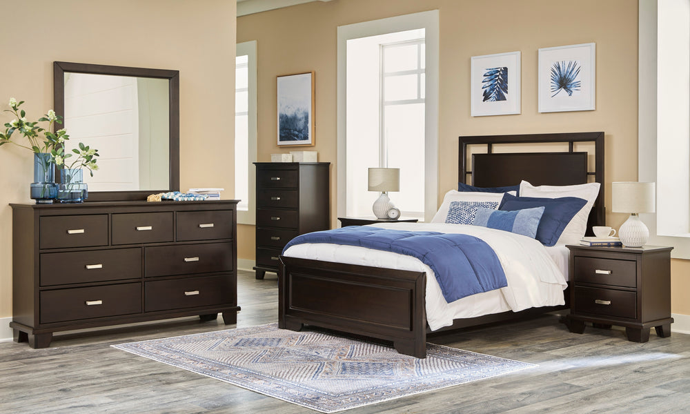 Conjunto de dormitorio con gavetero con espejo, cama y mesitas de noche de madera oscura en una habitación con grandes ventanas.