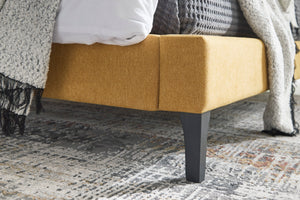 Cama amarilla con tela texturizada y patas oscuras sobre una alfombra estampada.
