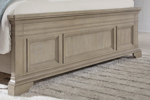 Pie de cama de estilo clásico en madera clara con paneles decorativos en relieve y patas robustas, ubicado sobre alfombra de textura suave en tonos neutros.