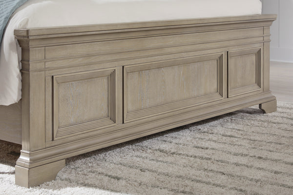 Pie de cama de estilo clásico en madera clara con paneles decorativos en relieve y patas robustas, ubicado sobre alfombra de textura suave en tonos neutros.