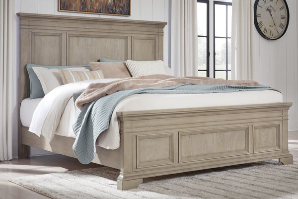 Cama matrimonial de estilo clásico en madera clara con cabecera y pie con paneles decorativos. Está vestida con ropa de cama en tonos neutros, incluyendo cojines y manta, y se muestra sobre fondo blanco.